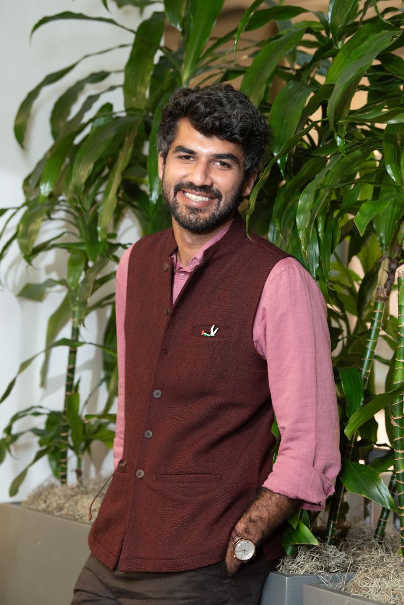 Man in maroon vest and pink shirt smiles, with greenery behind him.