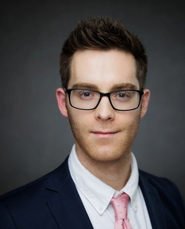 A man in a suit and tie, wearing glasses, looks directly at the camera.