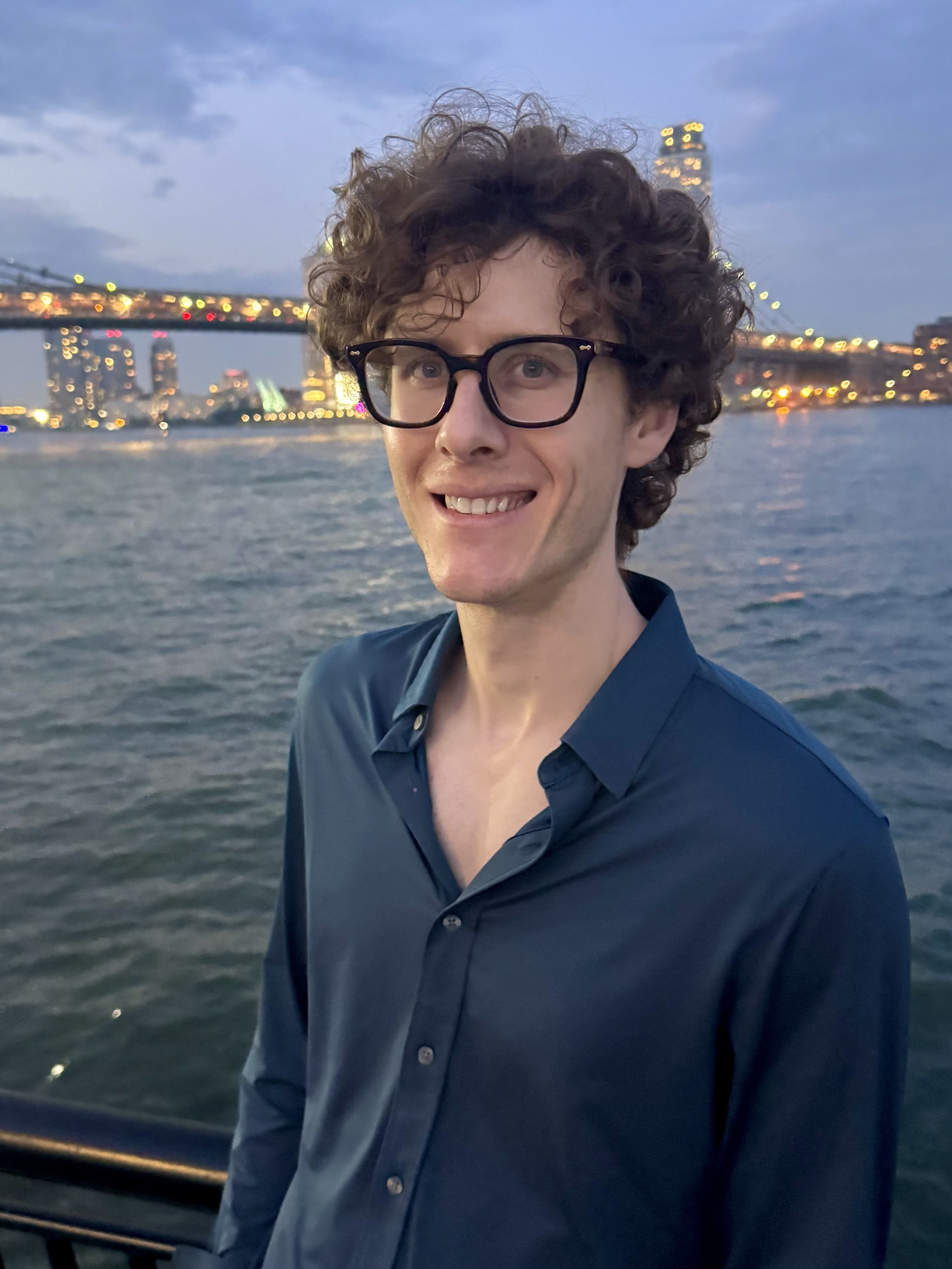 Young man with curly hair and glasses smiles by a waterfront with a bridge at dusk.