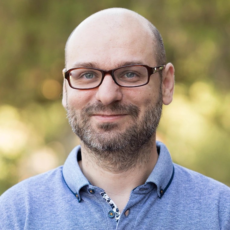 Bald man with glasses and a beard smiles, wearing a blue collared shirt.