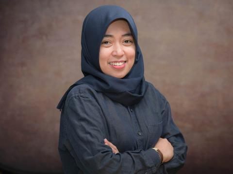 Nuzulul Putri, a smiling woman in a dark hijab and shirt poses with arms crossed.