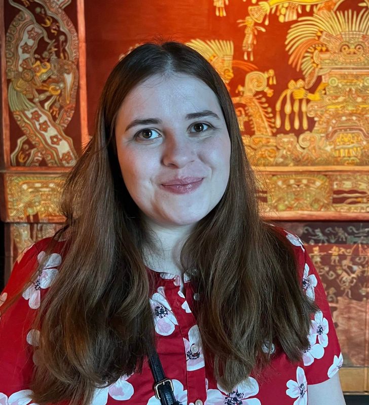 Young woman with auburn hair smiles at camera, wearing a red floral top.