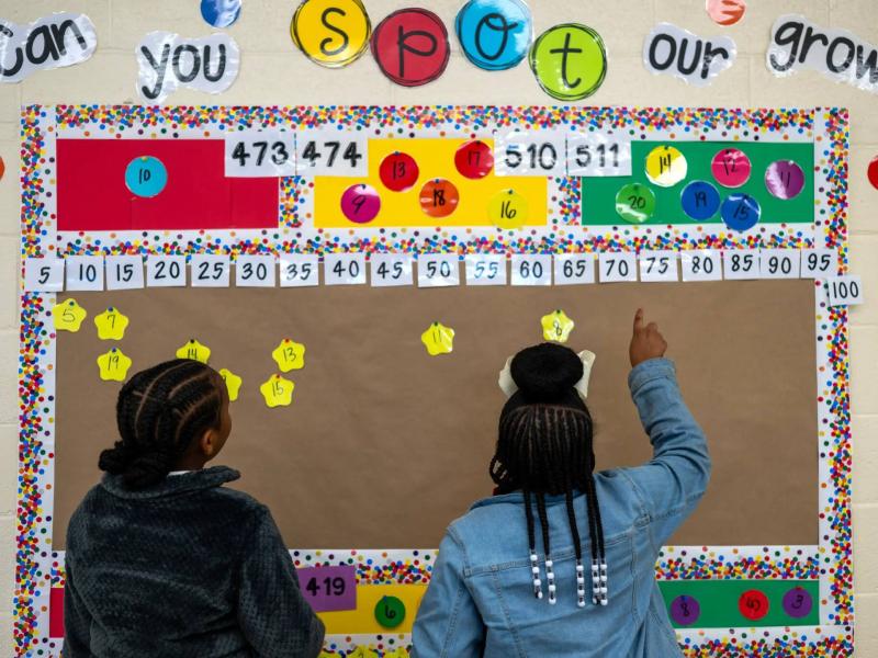 Two students point at a colorful classroom bulletin board displaying numbers.