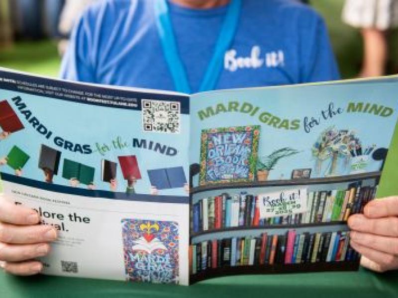 Person holds open colorful brochure about "Mardi Gras for the Mind" at a book fair.