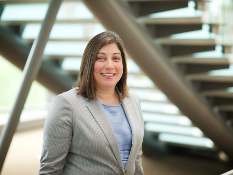 Woman in a light gray suit smiles at the camera in front of a modern staircase.