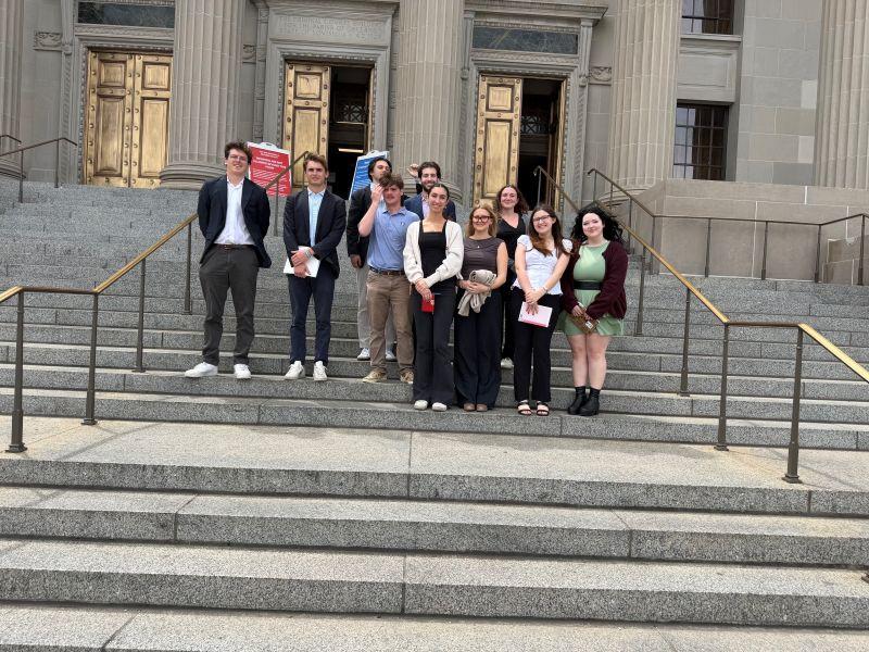 A group of ten young adults poses on the steps of a grand building.