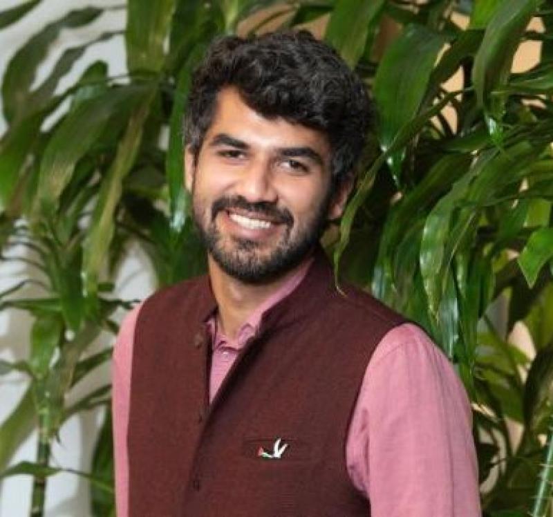 Man in maroon vest and pink shirt smiles, with greenery behind him.