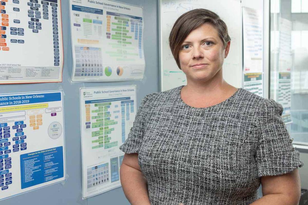 Woman with short brown hair in a grey tweed dress stands in front of a wall of charts.