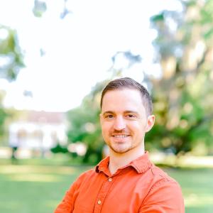 Man in orange shirt smiles with arms crossed outdoors.